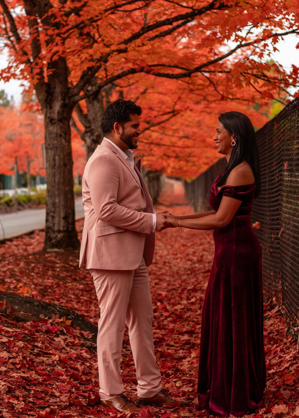 Couple holding hands under stunning red maple trees in fall, man in pink suit, woman in burgundy gown.