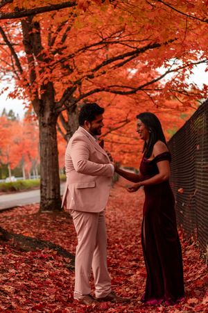 Couple smiling and holding hands beneath brilliant red autumn maple trees, dressed in elegant attire.