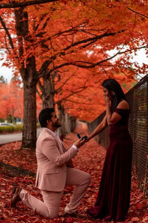 Man in pink suit proposes to woman in burgundy velvet dress amid stunning red autumn foliage.