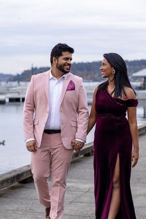 Engaged couple walking hand in hand along waterfront promenade in elegant autumn attire.