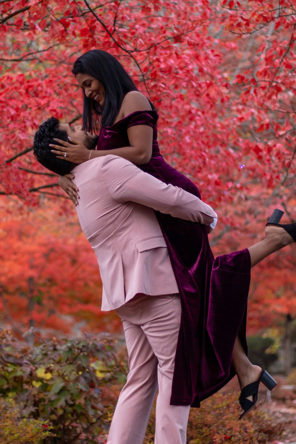 Joyful man lifting woman in burgundy velvet dress amid vibrant red autumn foliage.