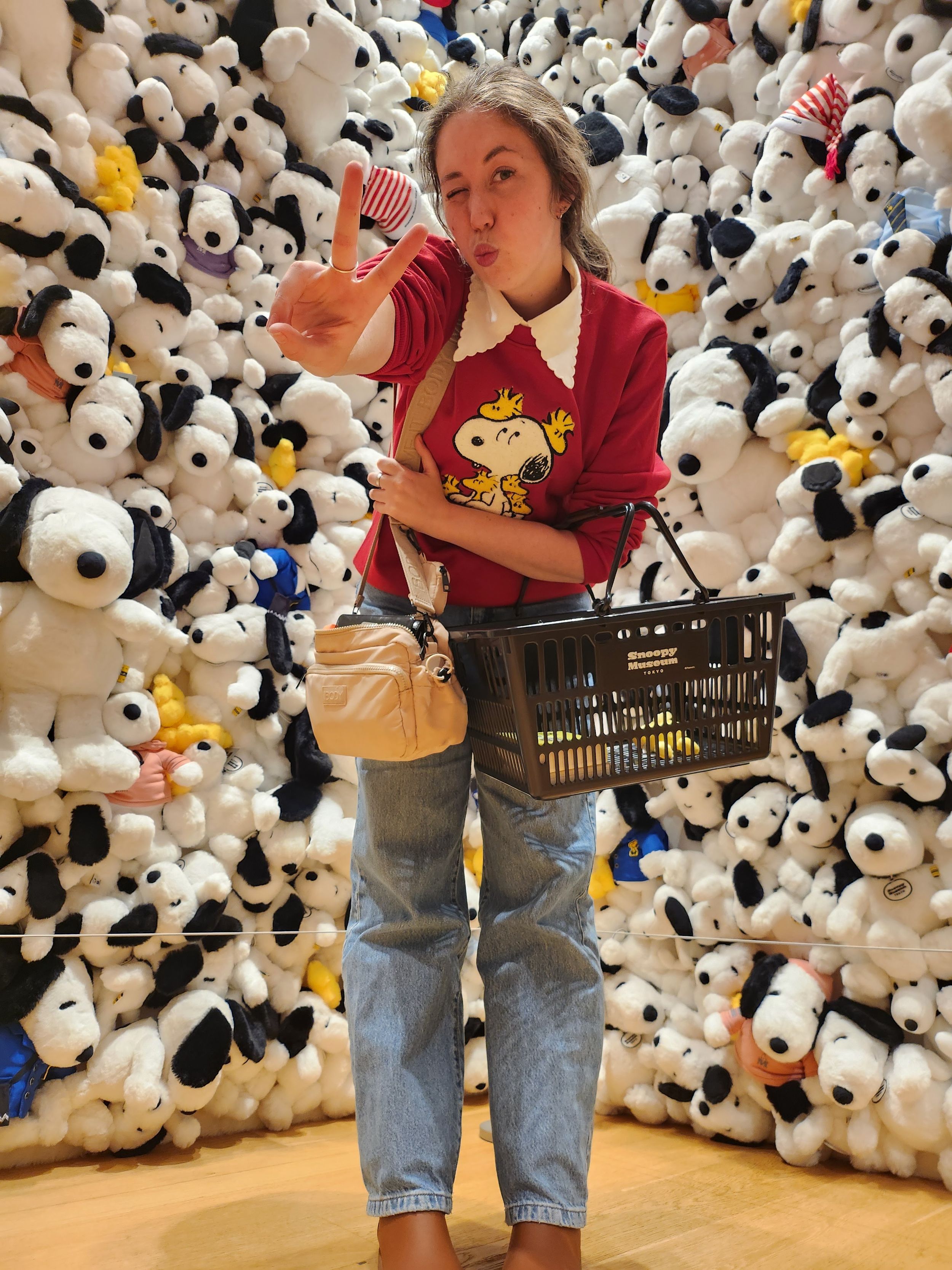 A person in jeans and burgundy shirt stands against wall of Snoopy plush toys at retail store display.