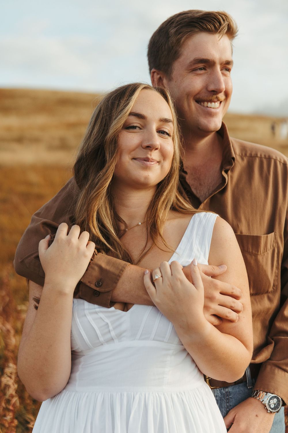 A romantic couple laughing and embracing in a golden field during sunset, wearing white and brown outfits.