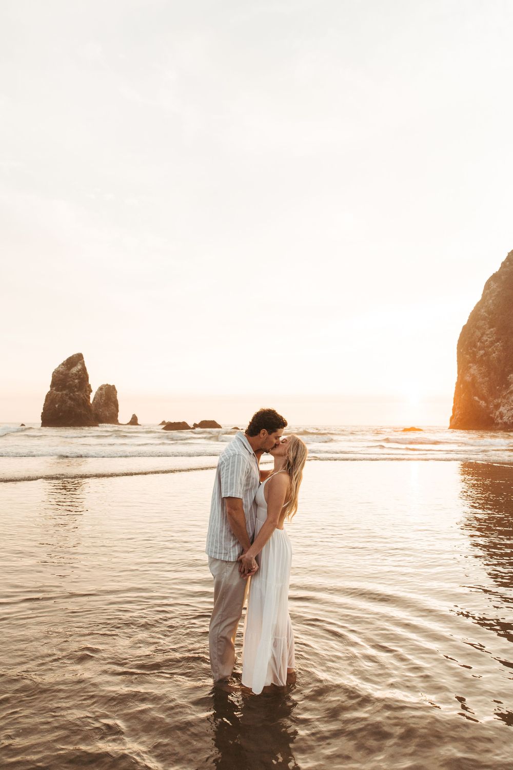 Couple standing in shallow ocean water at sunset near Cannon Beach's iconic Haystack Rock formation.