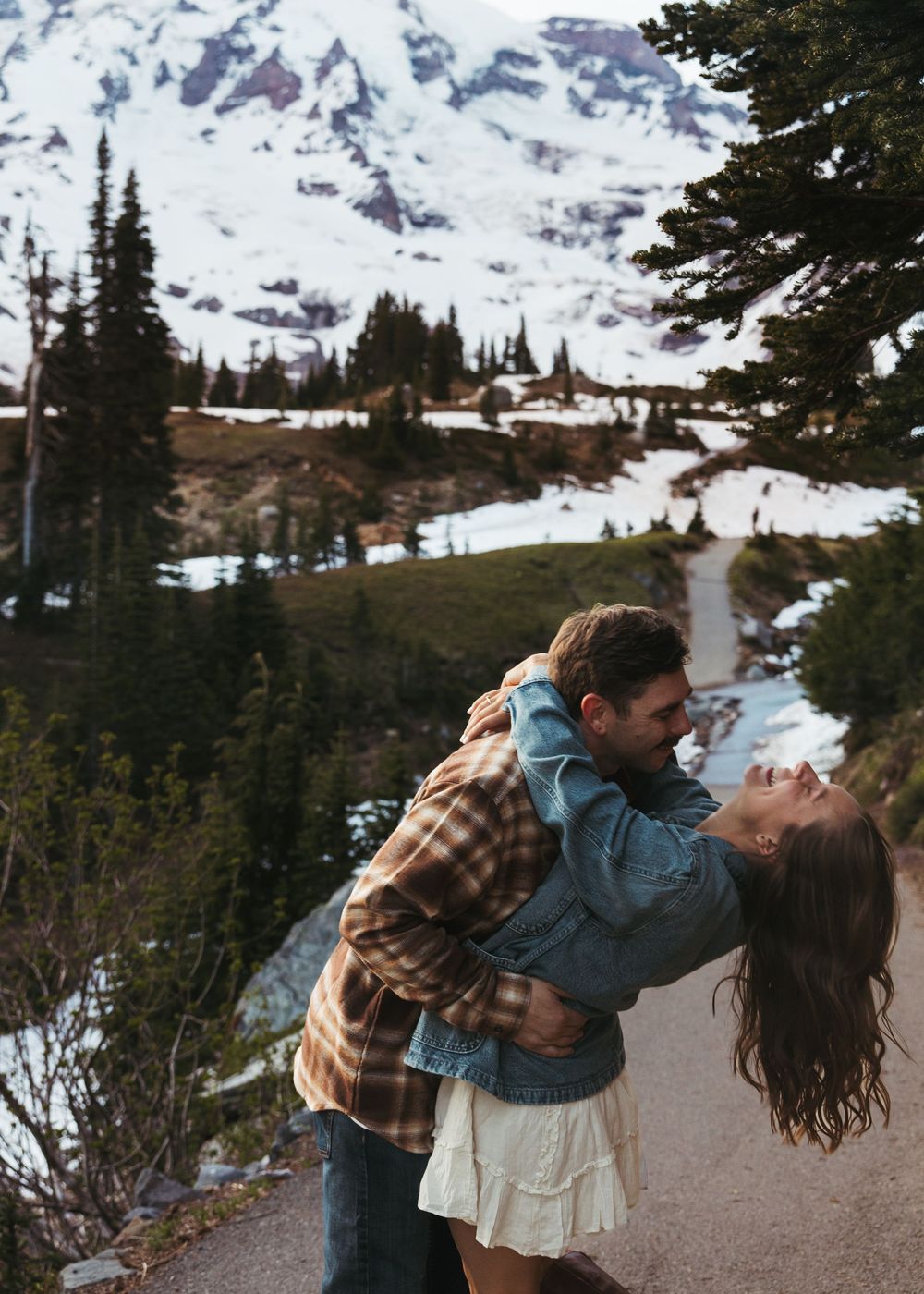 Couple sharing a romantic moment on a mountain trail with snow-capped peaks and evergreen trees in the background.
