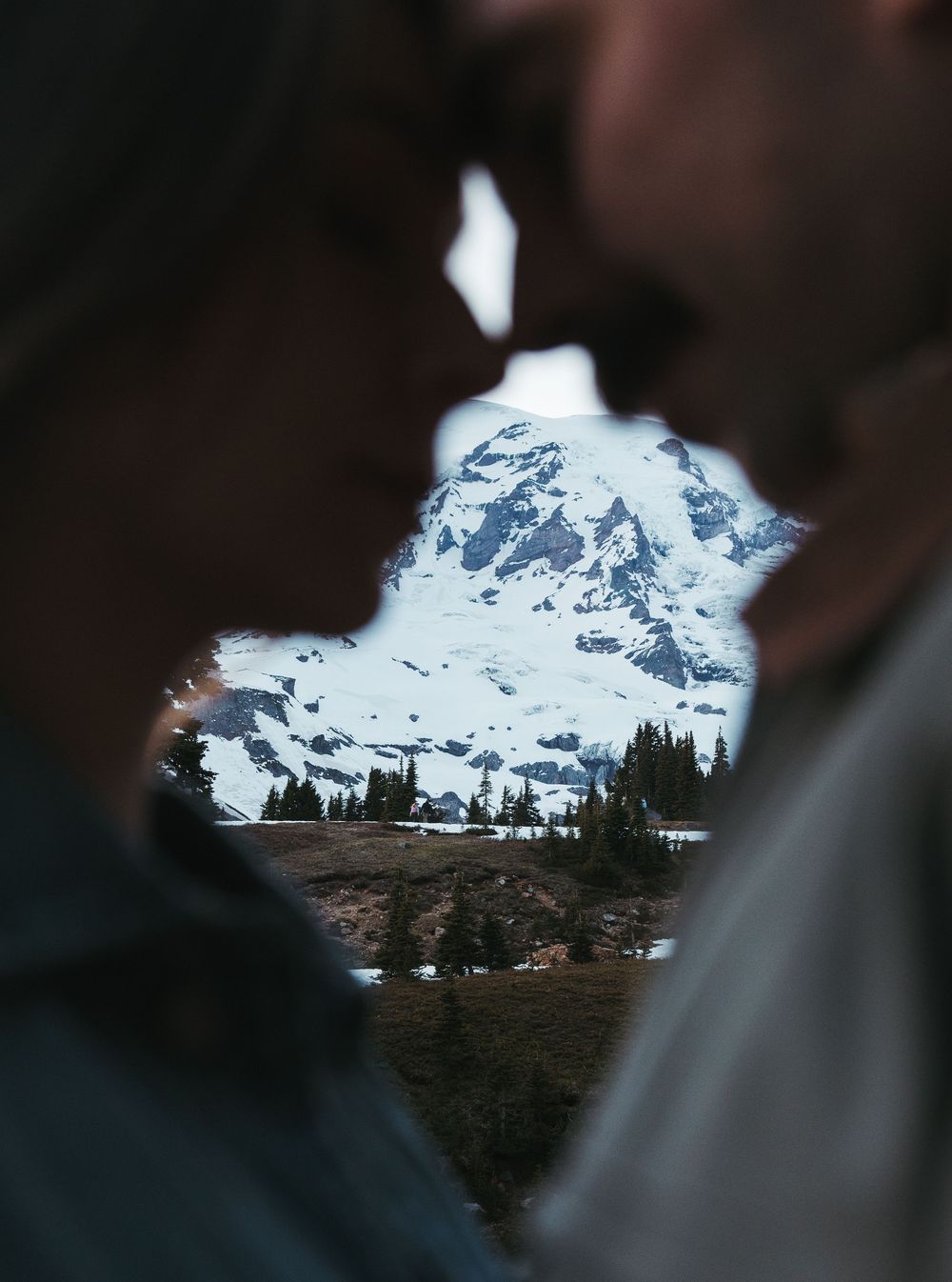 Couple sharing a kiss with a snow-capped mountain and alpine lake visible between them in soft focus.