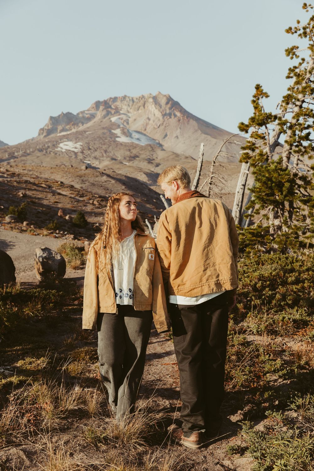 A couple in warm outdoor clothing stands together against a scenic mountain landscape at sunset.