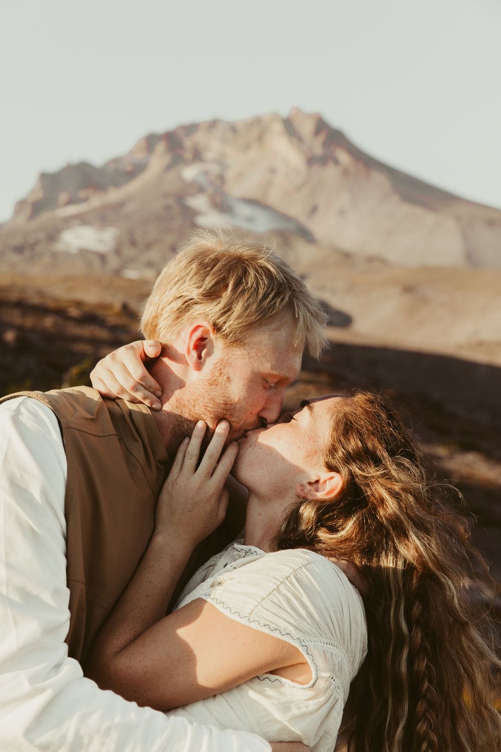 A couple shares romantic kisses against a backdrop of rugged desert mountains at sunset.