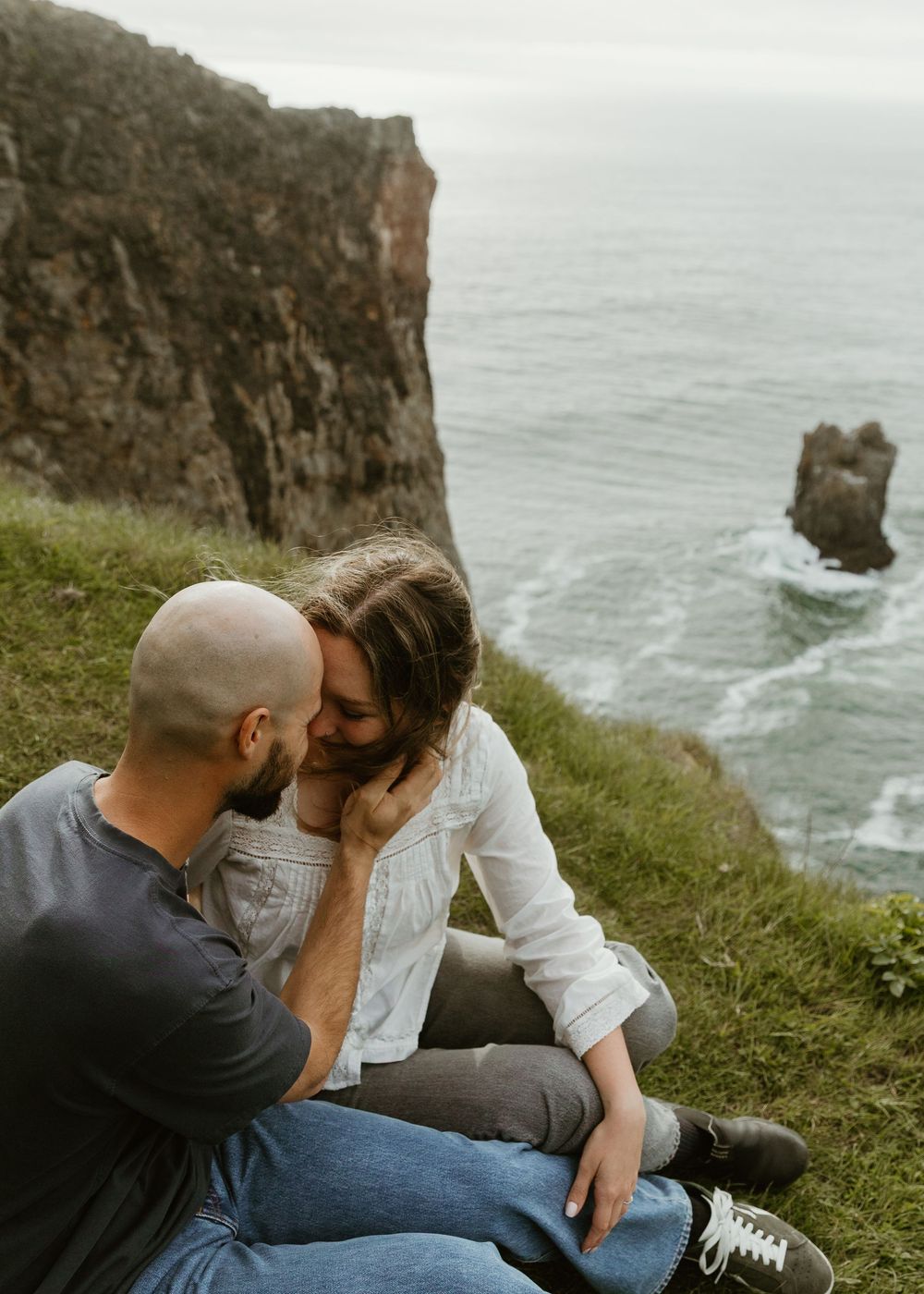 Couple sitting on a grassy cliff edge sharing a romantic moment with ocean rocks and sea in the background.