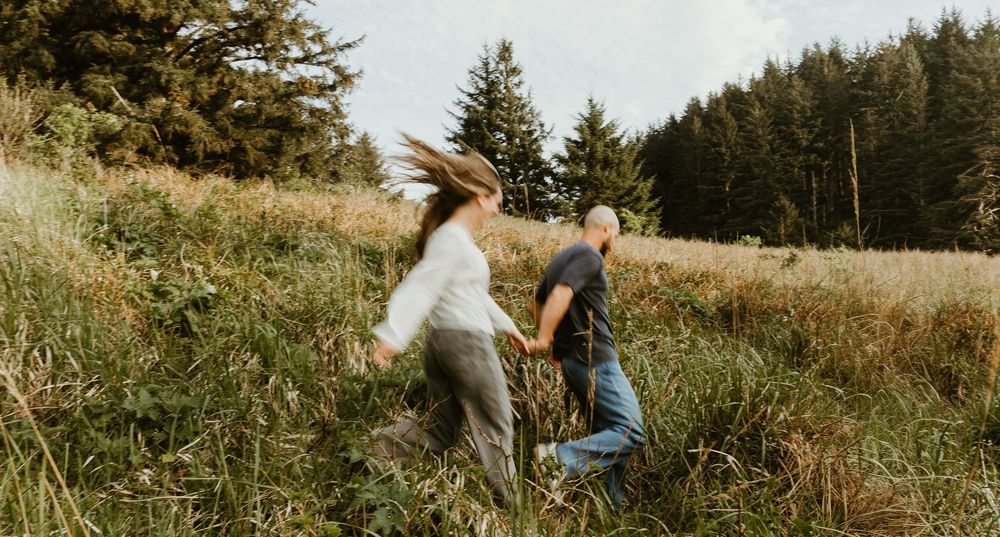 Couple holding hands running through a lush green meadow with tall pine trees in the background.