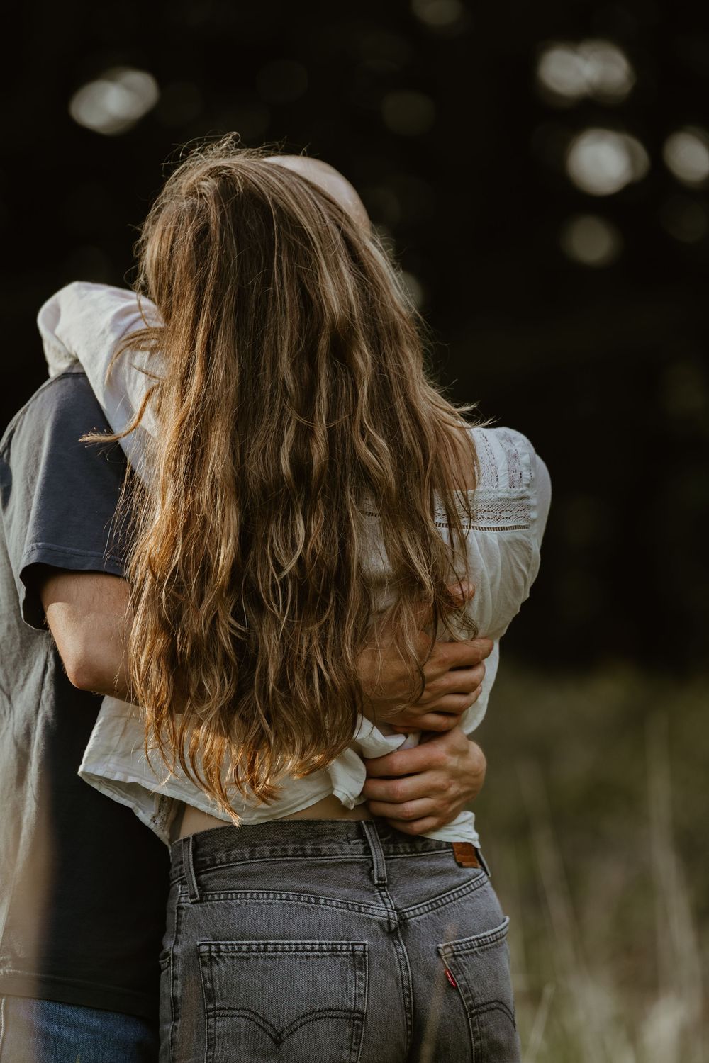 Couple embracing from behind outdoors, woman with long wavy hair wearing gray Levi jeans in a natural setting.