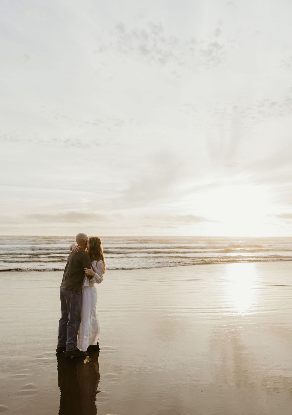 Couple embracing on a reflective beach at sunset, waves gently rolling in behind them.