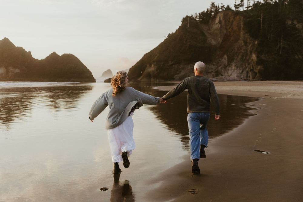 Elderly couple holding hands running joyfully on a scenic beach with rocky cliffs and calm water in the background.