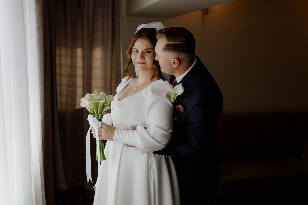 A bride in a white winter wrap stands with her groom in an elegant indoor setting during their winter wedding.