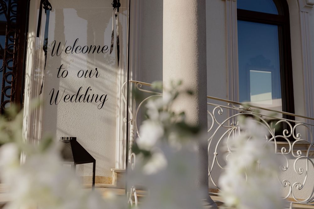 Elegant wedding welcome sign with white text displayed in an ornate frame against a light colored wall.