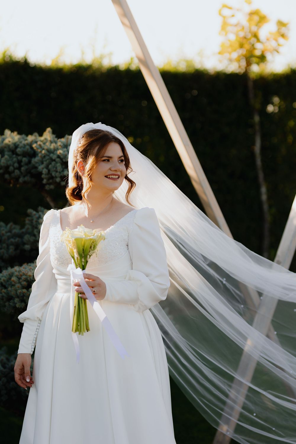 A bride in a white wedding dress stands outdoors with a flowing veil catching the sunlight in a romantic setting.