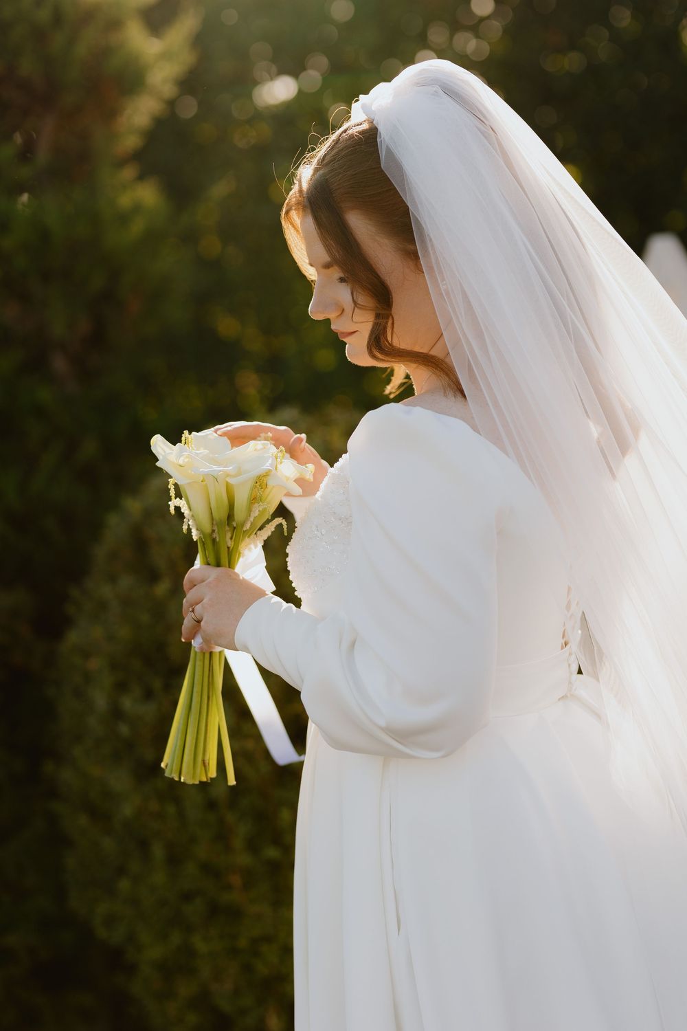 Bride in long-sleeved white dress holds a bouquet of roses and calla lilies in golden sunlight.