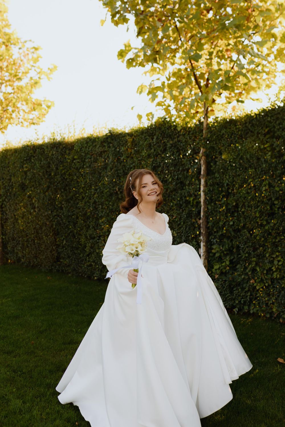 Bride in flowing white dress twirling joyfully in garden with golden sunlight filtering through trees.