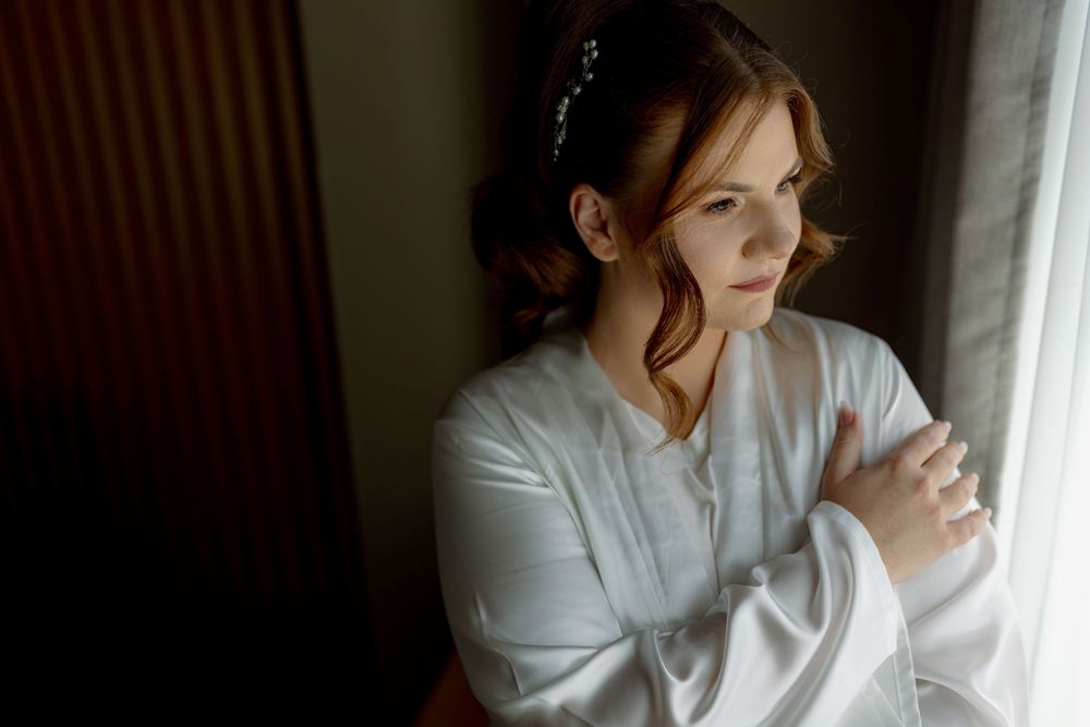 Woman in a light gray sweater sits by window holding a coffee mug while gazing thoughtfully outside.