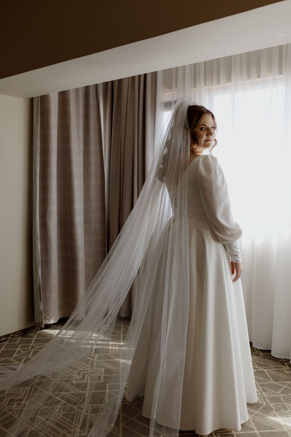 Elegant bride in a long-sleeved white wedding dress and flowing veil poses by a window with sheer curtains.
