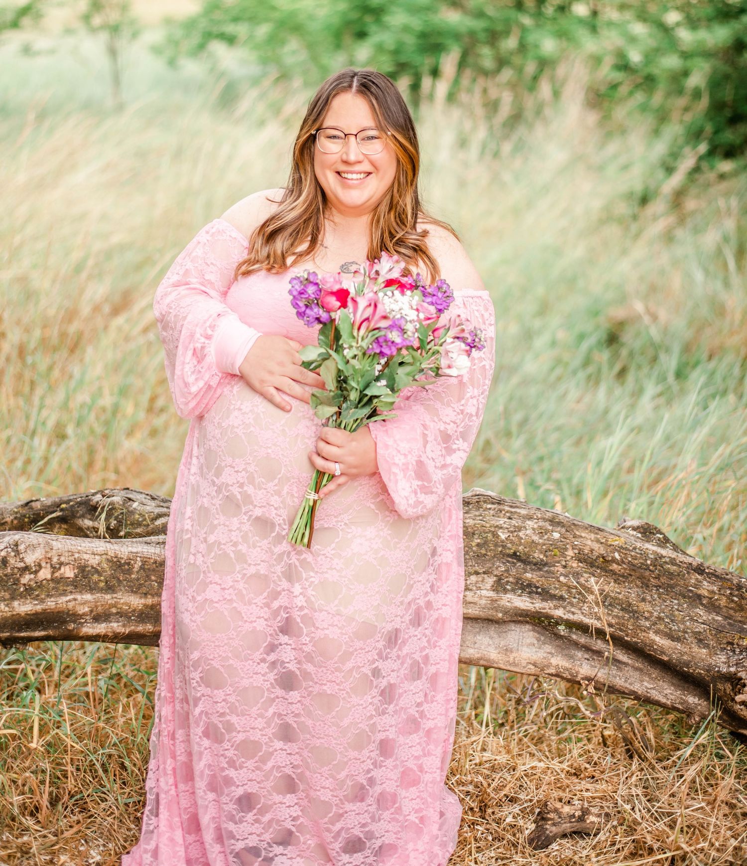 Someone in a pink lace dress holds a vibrant bouquet of flowers while standing in a field of tall grass.