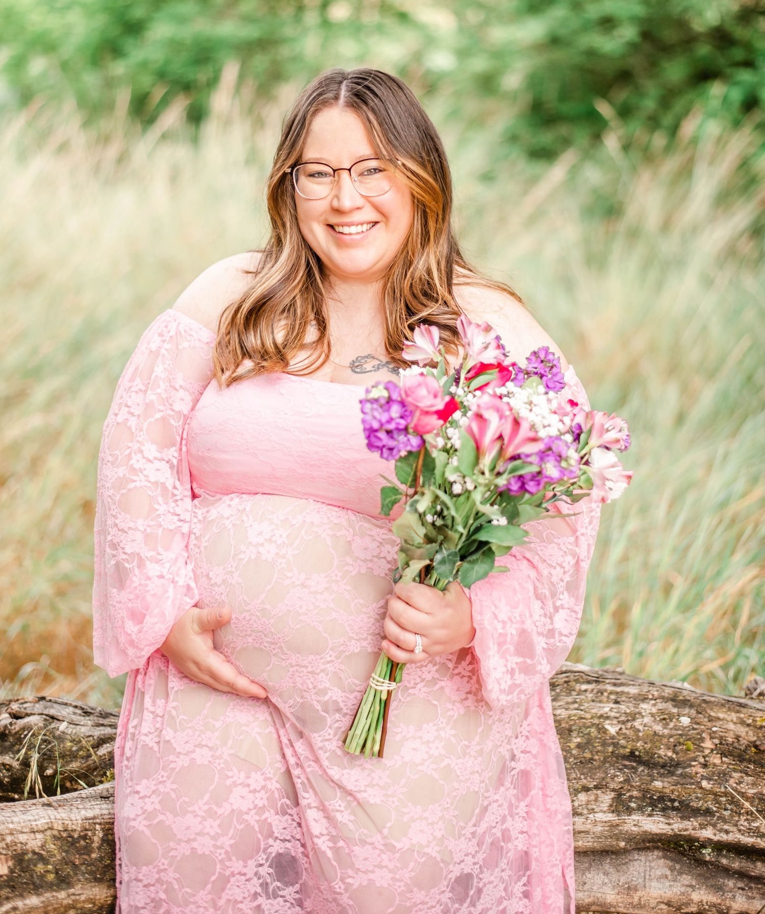 A person in a pink lace dress poses outdoors holding a bouquet of wildflowers in a natural setting.
