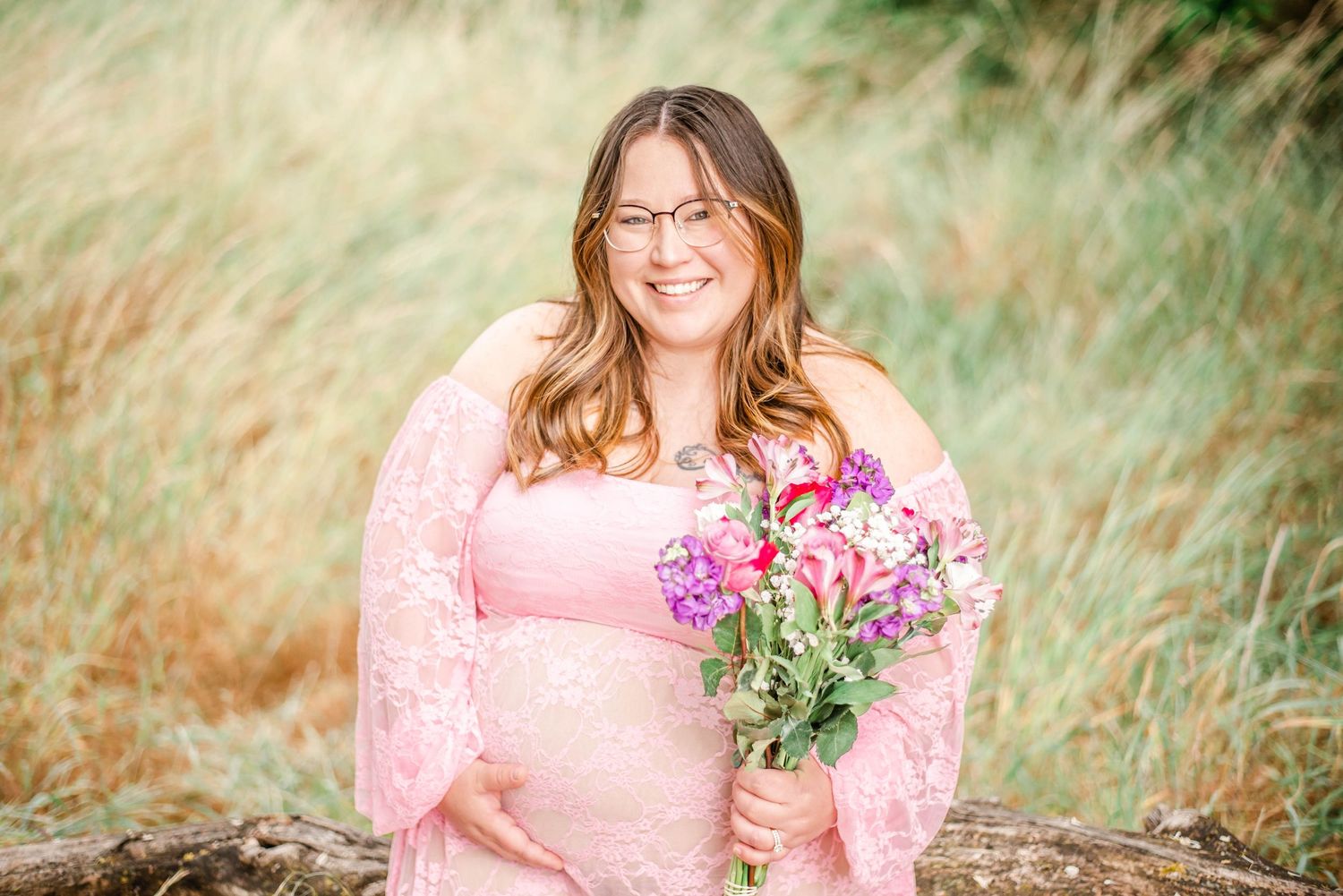 A person wearing a pink sweater sits in tall grass holding a bouquet of wildflowers during a golden hour photoshoot.