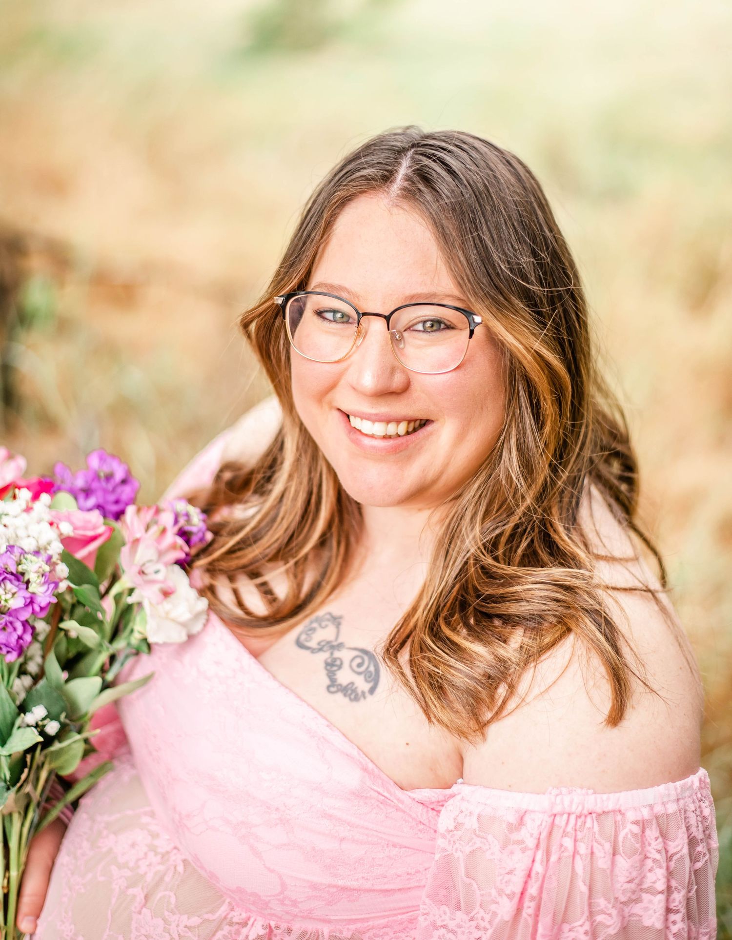 A portrait of someone wearing glasses and a pink dress holding a bouquet of purple and pink flowers outdoors.