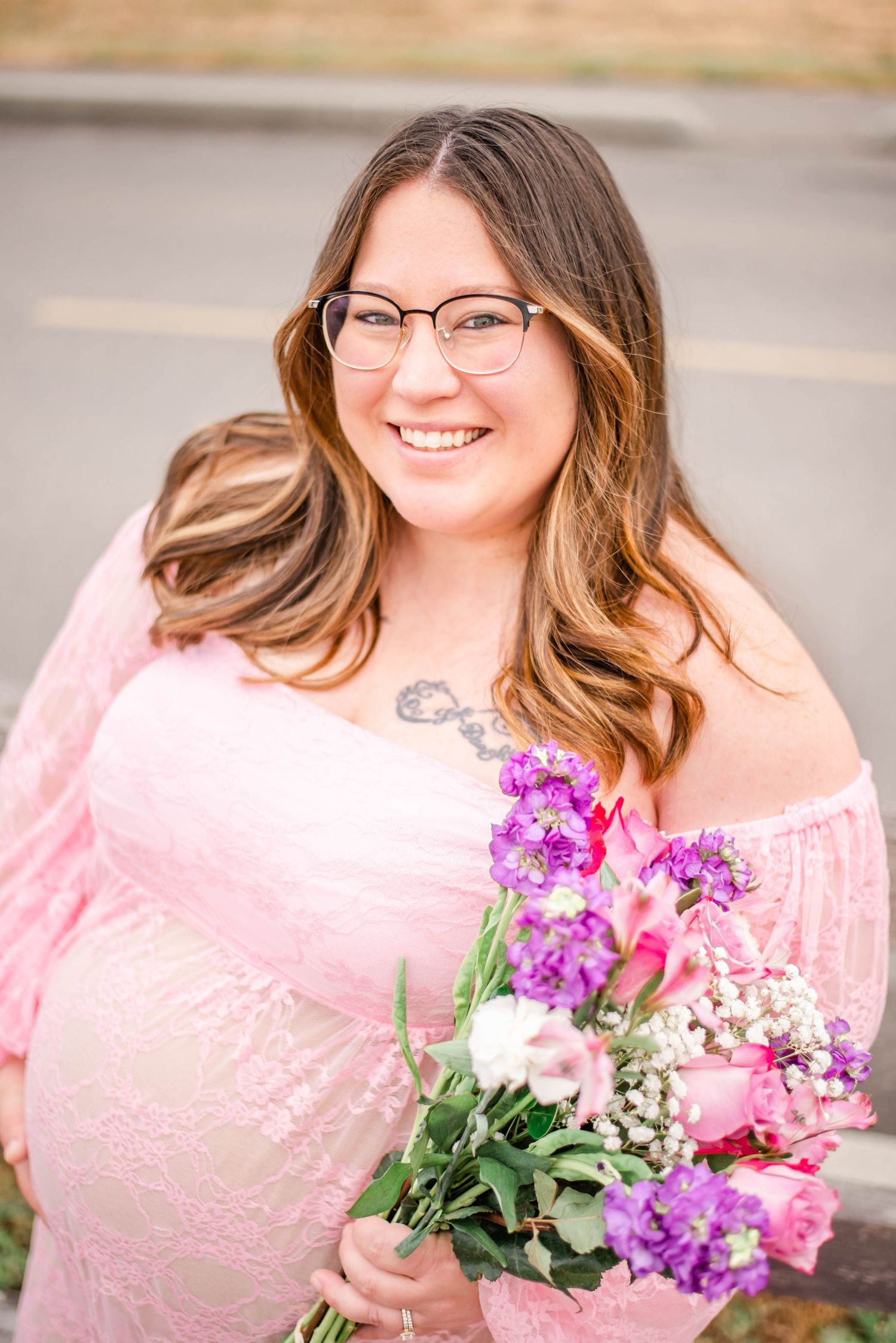 Expectant mother in pink lace dress holding colorful bouquet of purple and pink flowers outdoors.