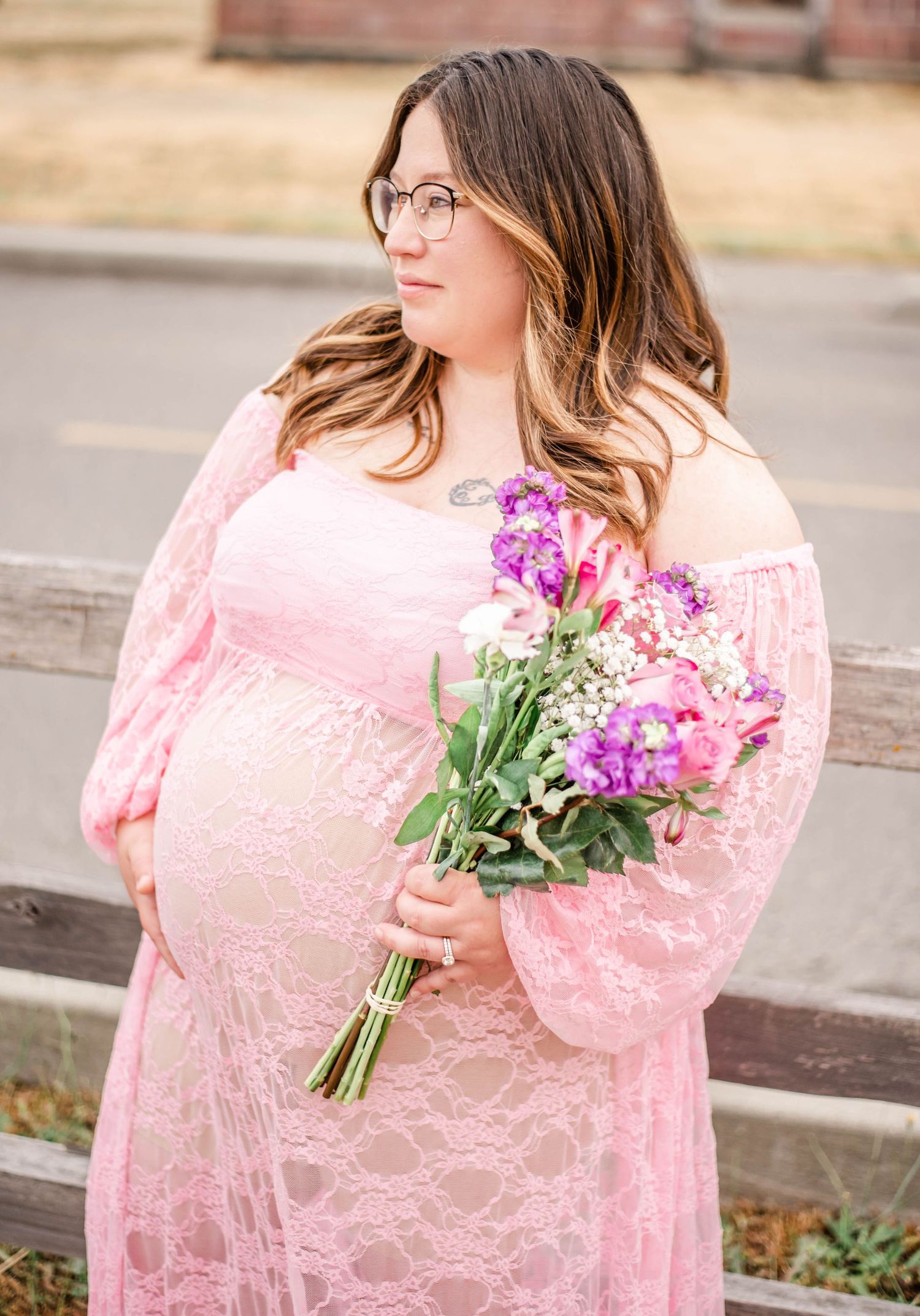 Urban photoshoot with pink lace dress and purple floral bouquet against brick wall and sidewalk backdrop.
