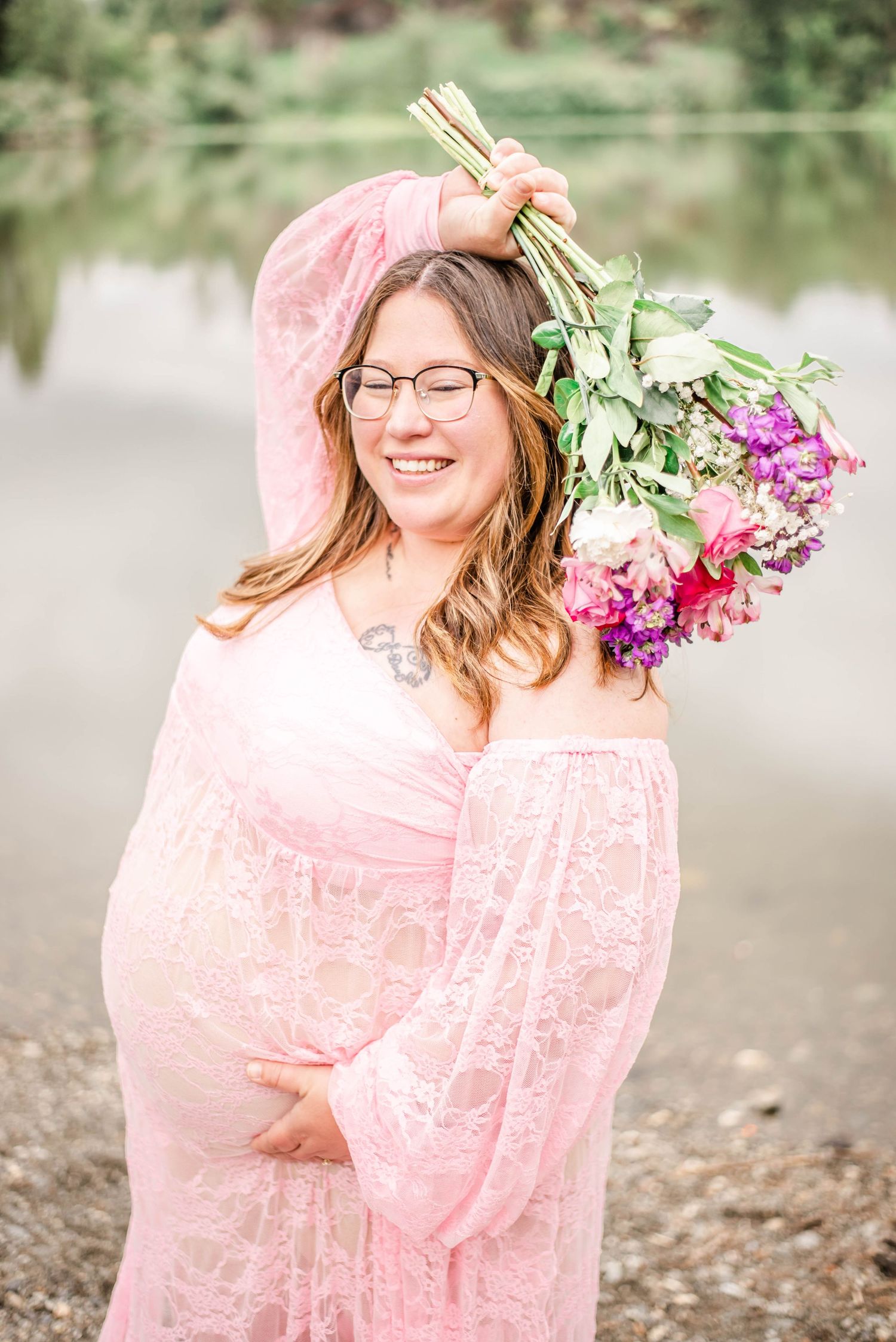 Romantic maternity photoshoot by a lake featuring a flowing pink lace dress and purple floral bouquet.