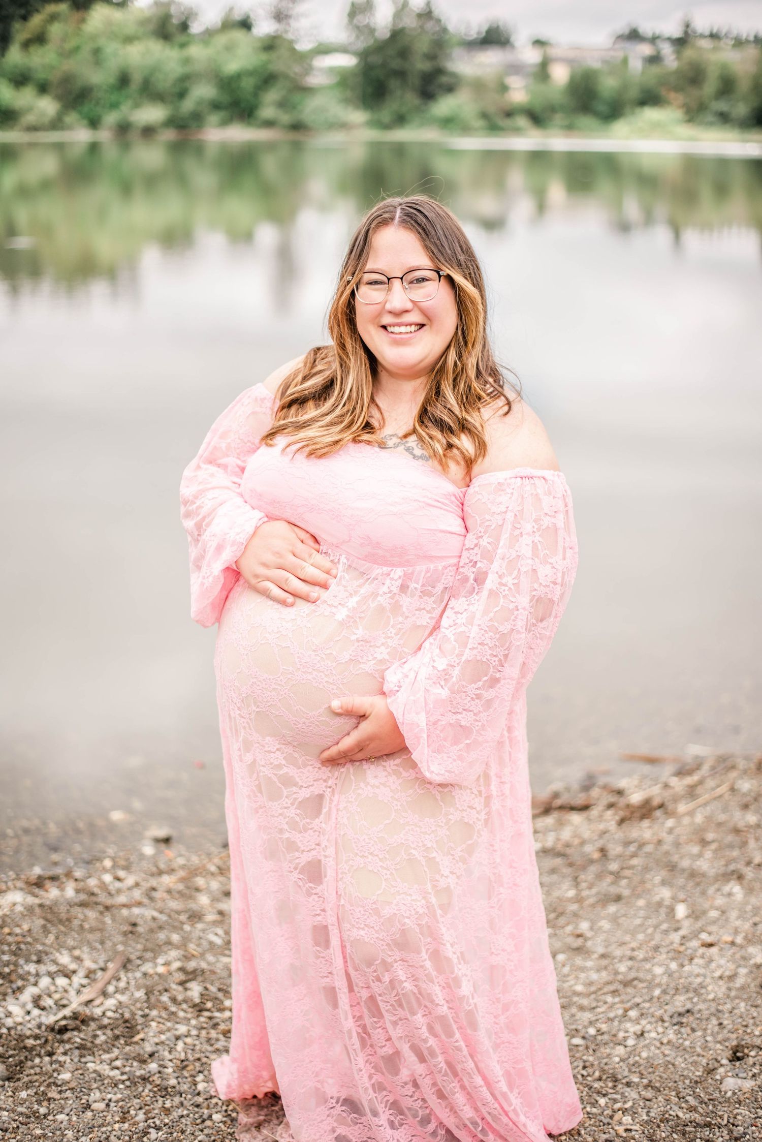 Outdoor maternity session by waterfront with soft pink lace gown and natural lighting.