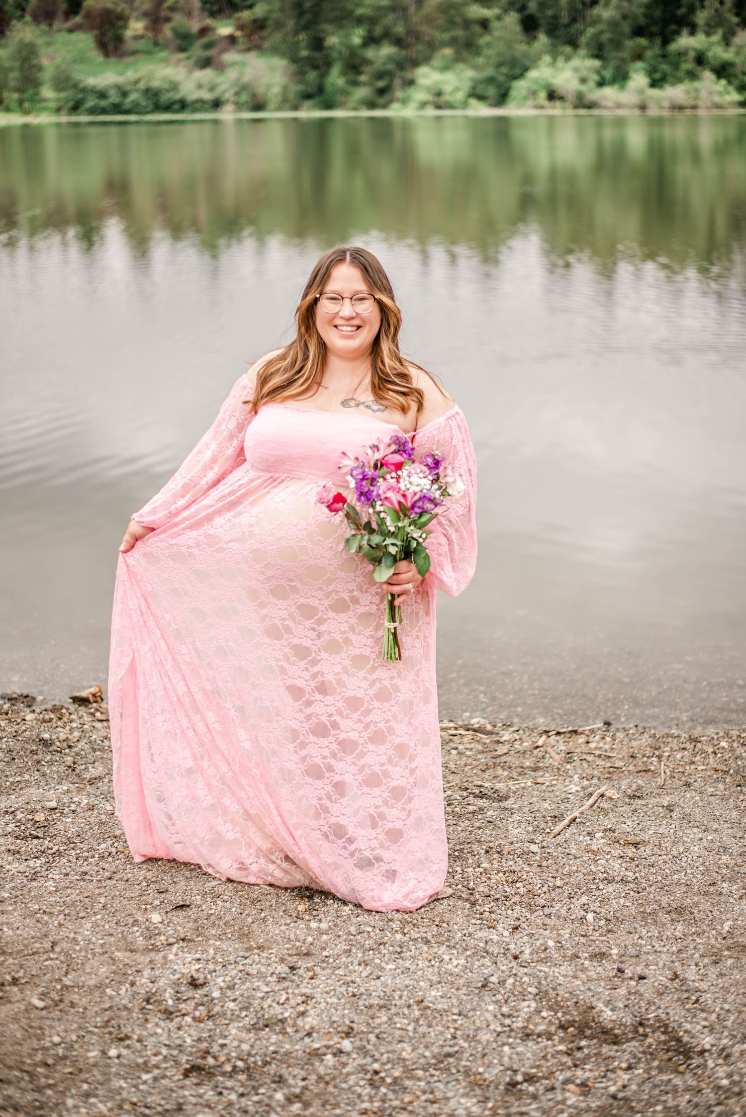 A romantic lakeside portrait in a flowing pink lace gown holding a vibrant purple and pink bouquet.