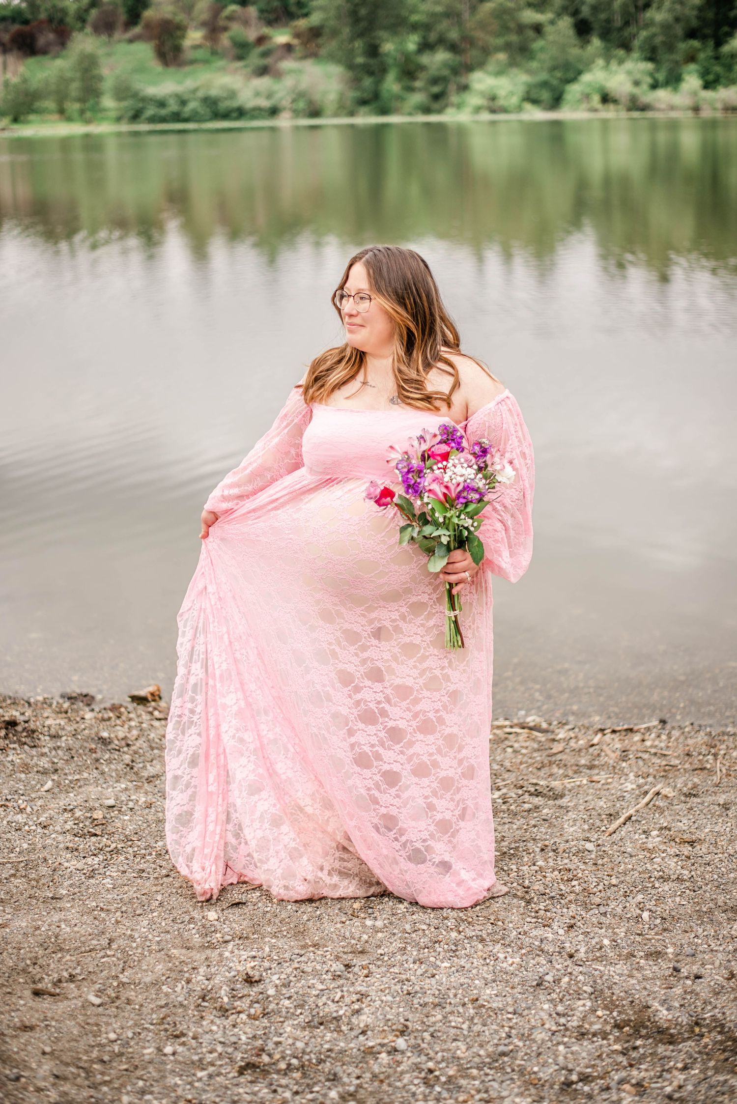 A person in a flowing pink lace dress sits gracefully by a serene lake on a rocky shore.