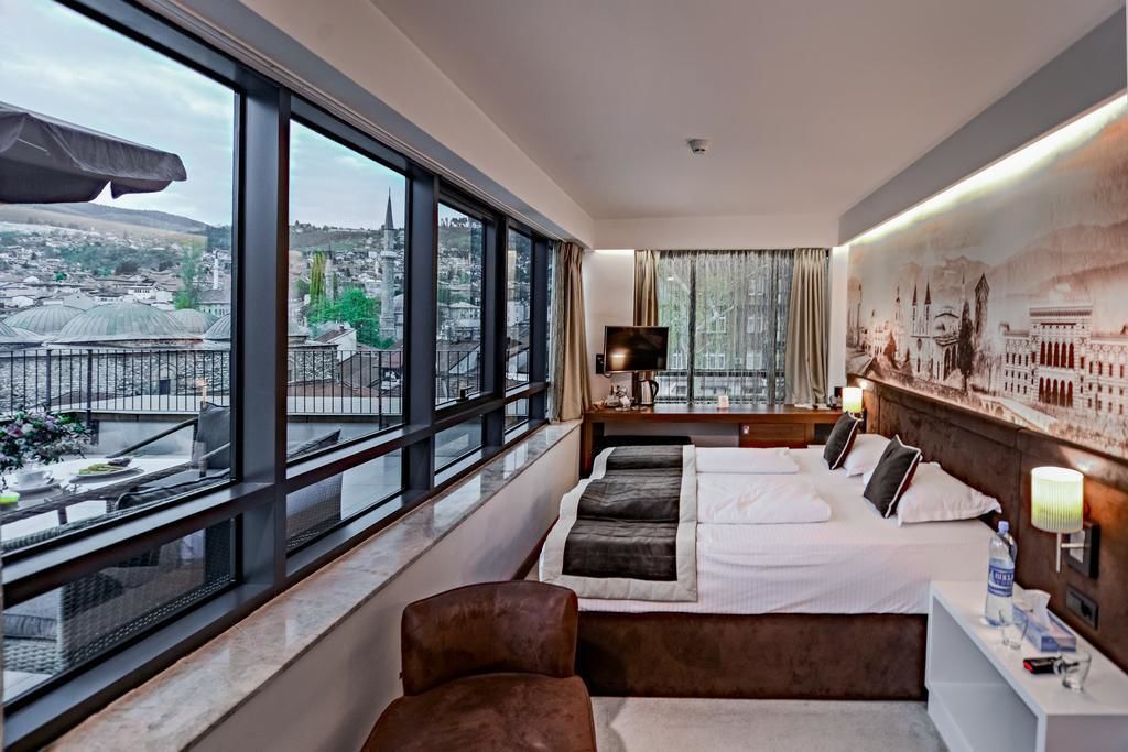 Modern hotel room with panoramic city views featuring white bedding, brown accents, and a decorative wall mural.