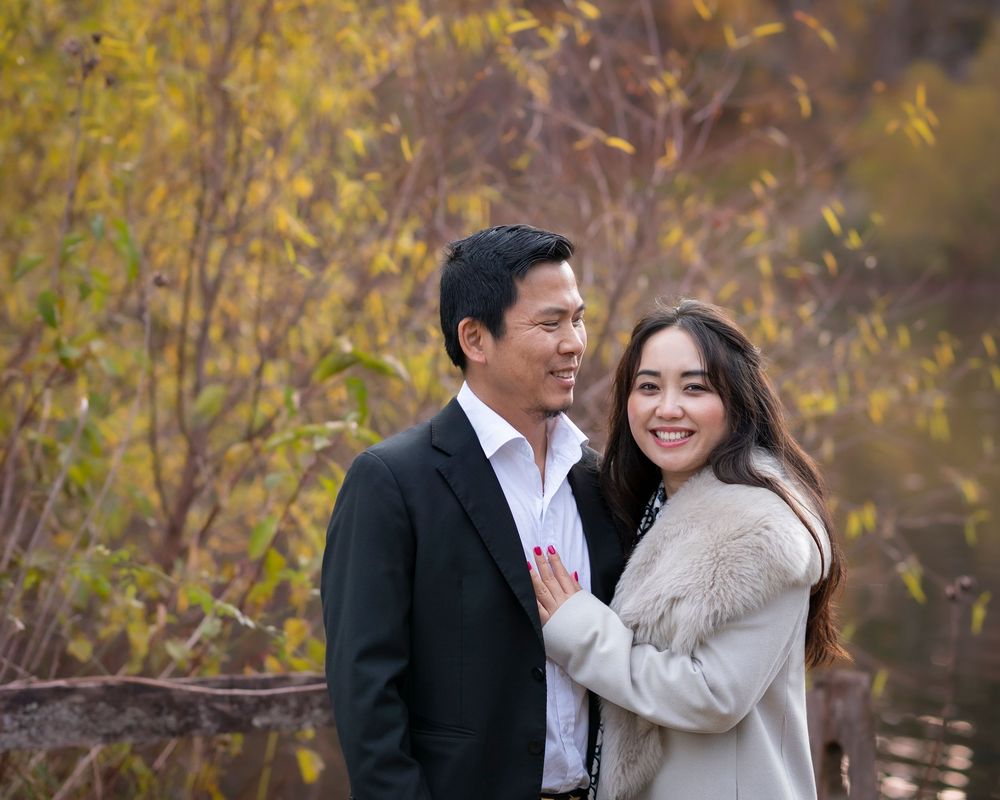 Couple posing together outdoors in autumn, man in black suit and woman in gray coat, smiling warmly. Holmdel Park NJ