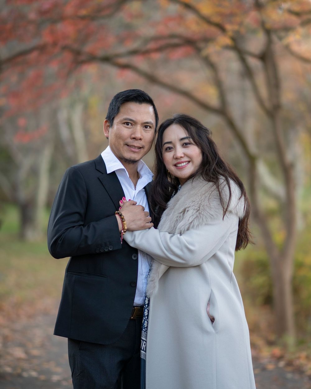 Couple embracing outdoors in autumn park, man in dark suit, woman in cream coat, colorful fall foliage background. Monmouth County NJ