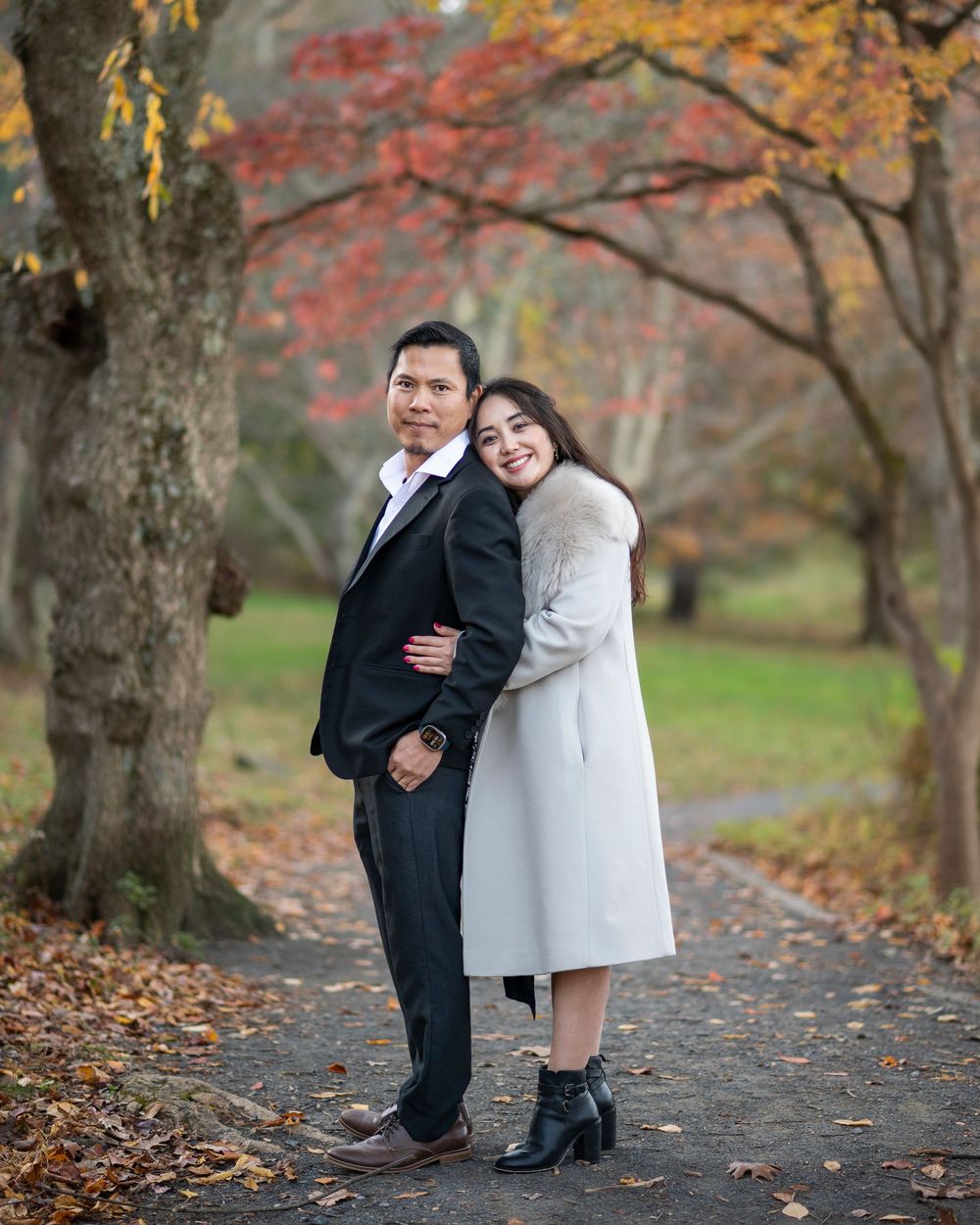 Couple posing together on autumn park path surrounded by colorful fall foliage, woman in white coat leaning on mans shoulder. Holmdel Park