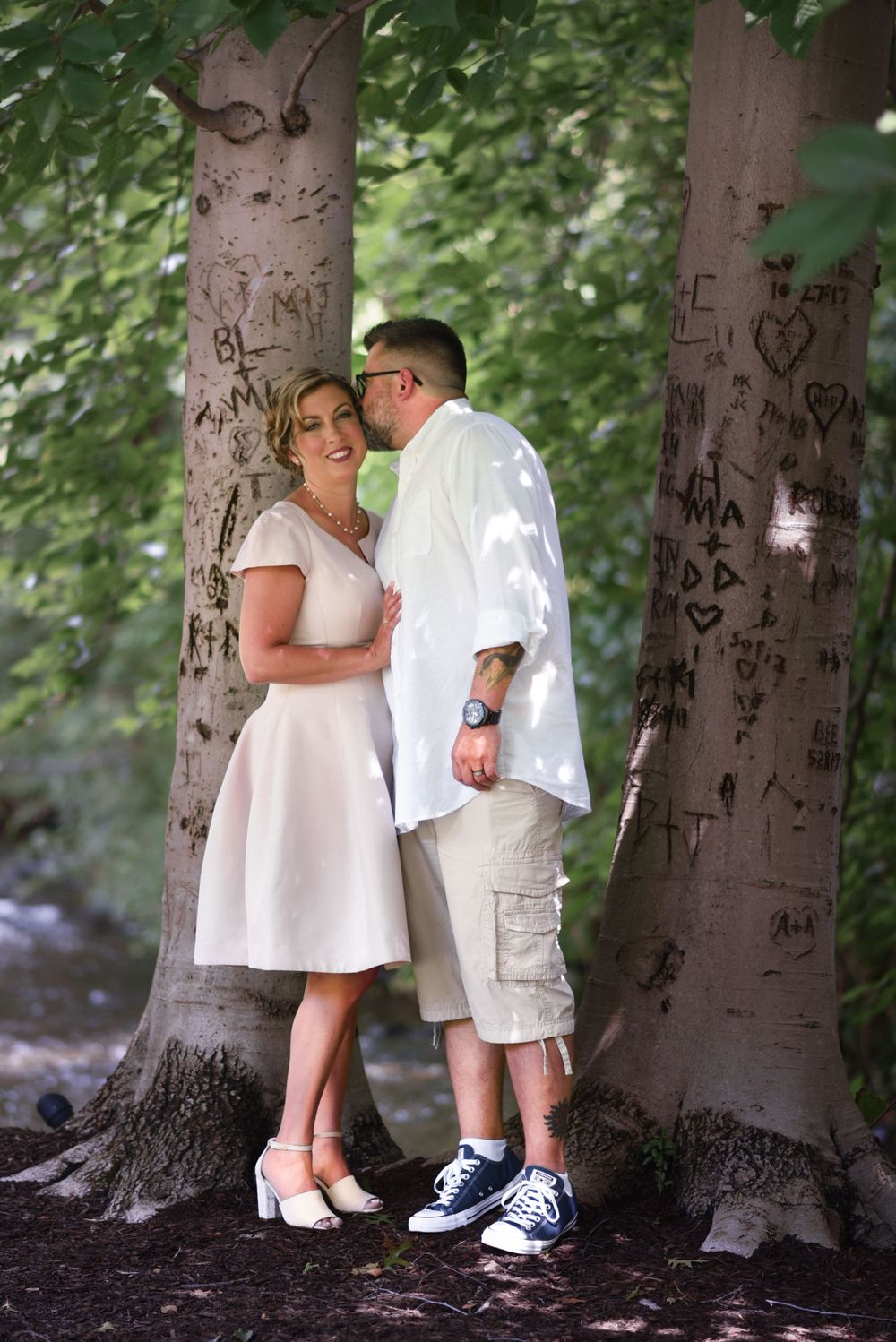 A portrait of a Couple in white embracing near tree trunks covered in carved initials and hearts.Middletown NJ