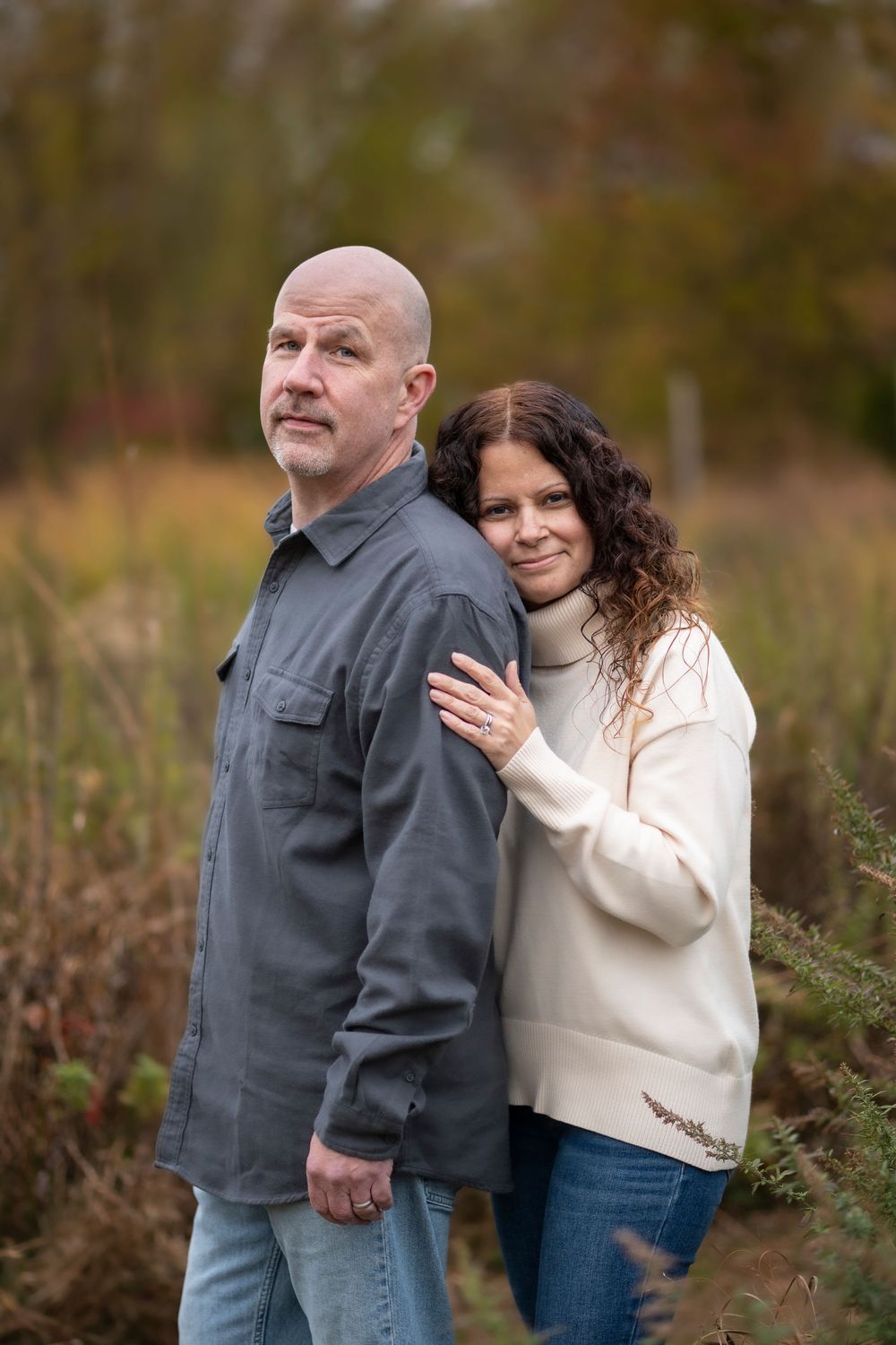 Couple posing back-to-back in an autumn field, woman leaning on mans shoulder smiling warmly.Poricy Park Middletown NJ Monnouth County