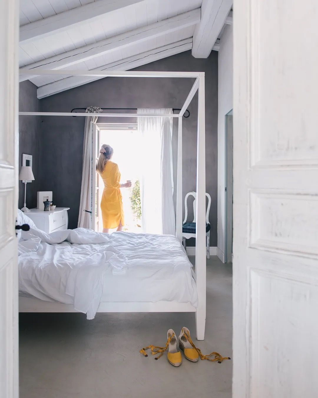 A woman in a yellow dress steps into a bright, cozy bedroom with white walls and soft natural light.