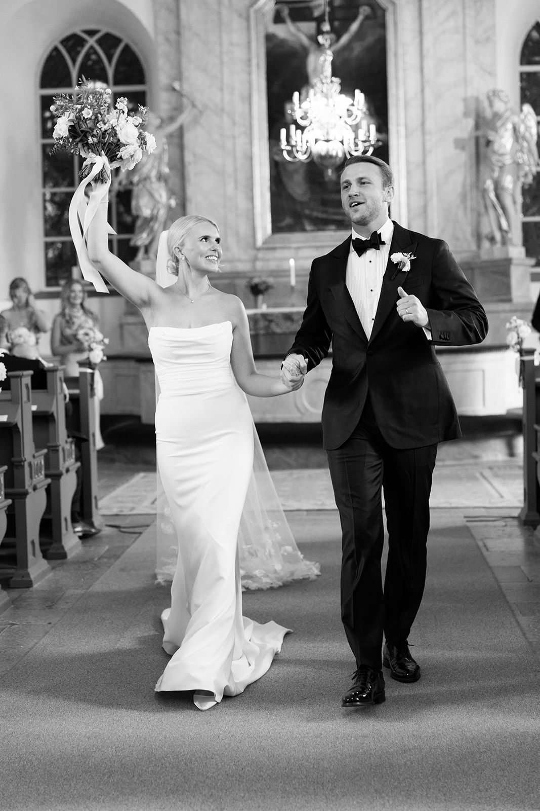 A bride and groom dance together in an elegant venue with arched windows and columns in black and white.