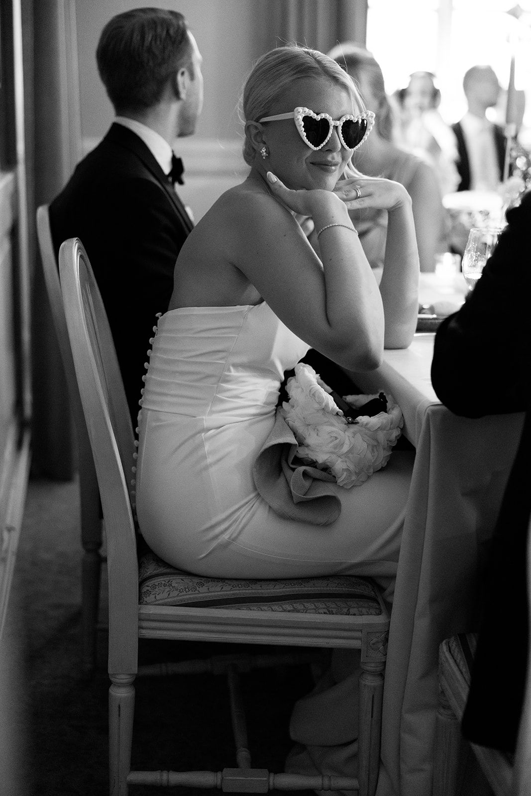 A person in a white dress and heart-shaped sunglasses sits at a table during what appears to be a formal event.