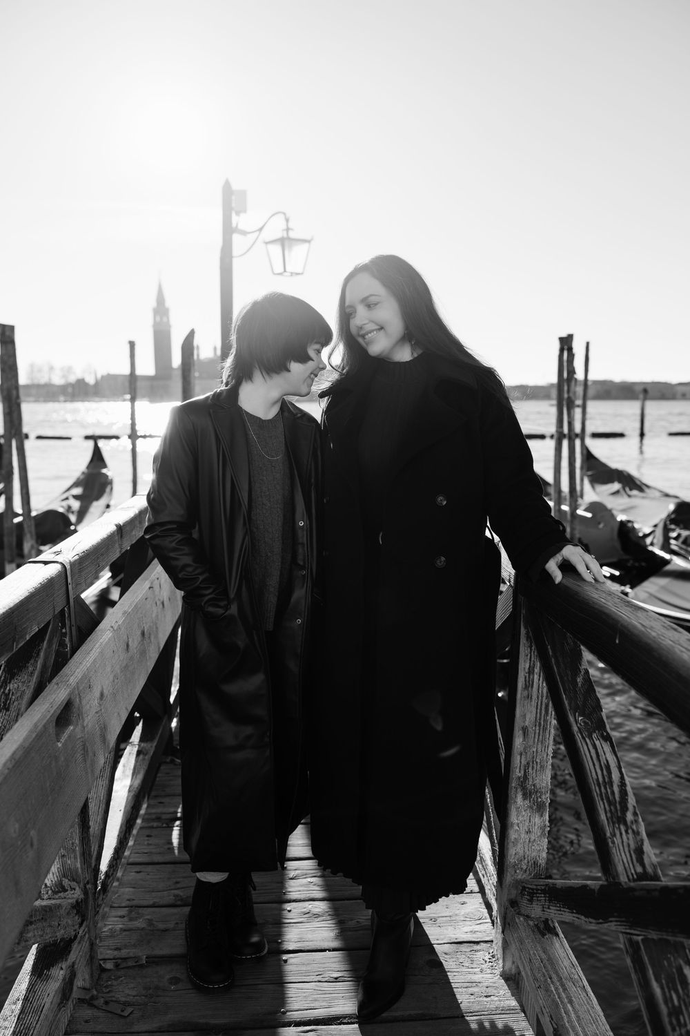 A black and white photo of two people in dark coats standing together on a wooden dock in Venice with gondolas visible.