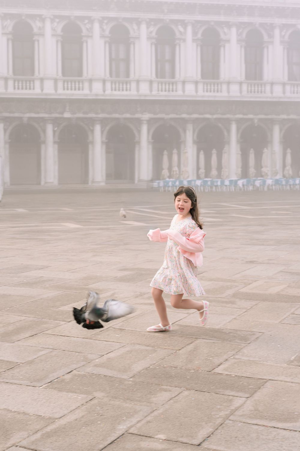 A young girl in a pink dress dances playfully in St. Mark's Square in Venice while a pigeon flies nearby.