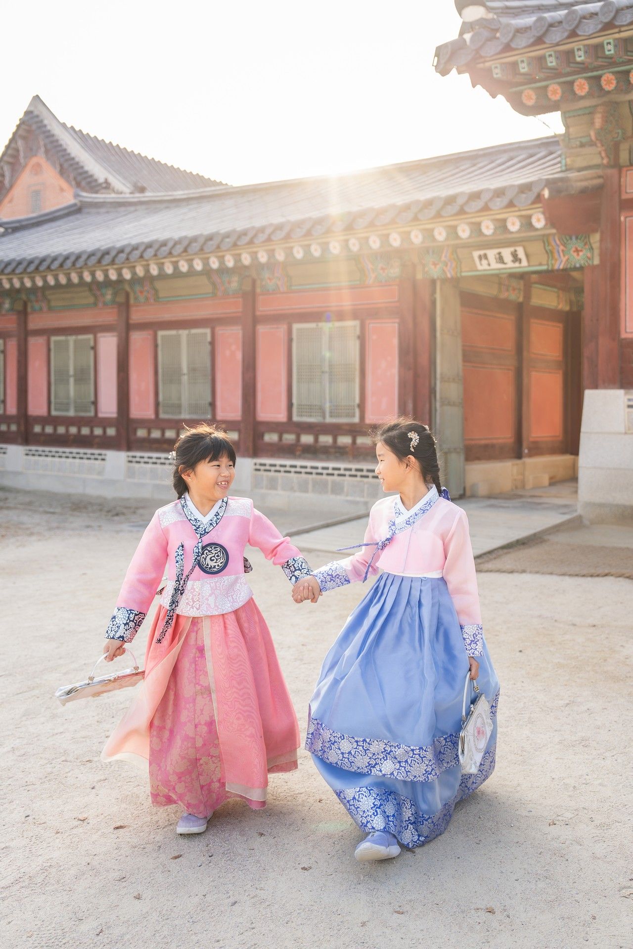 Two people in traditional Korean hanbok dresses walk together in a historic palace courtyard with traditional architecture.