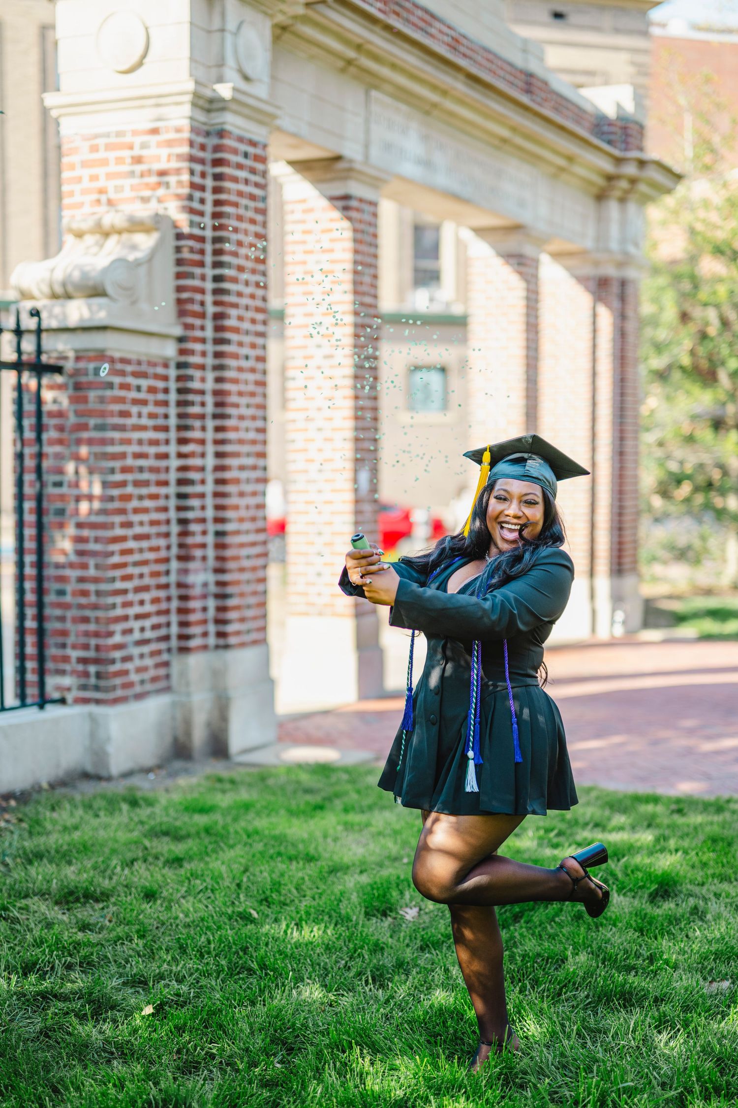 A graduate in a dark green cap and gown poses joyfully on a grassy lawn in front of a brick building.