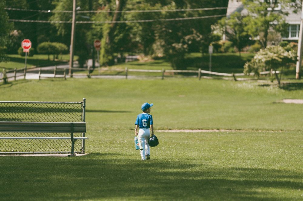 Young baseball player in teal uniform walking across green outfield grass during a little league game.
