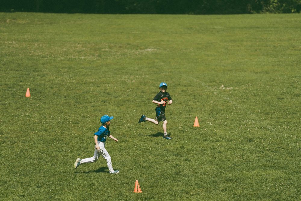 Two young baseball players in teal uniforms running drills with orange cones on a green grass field.