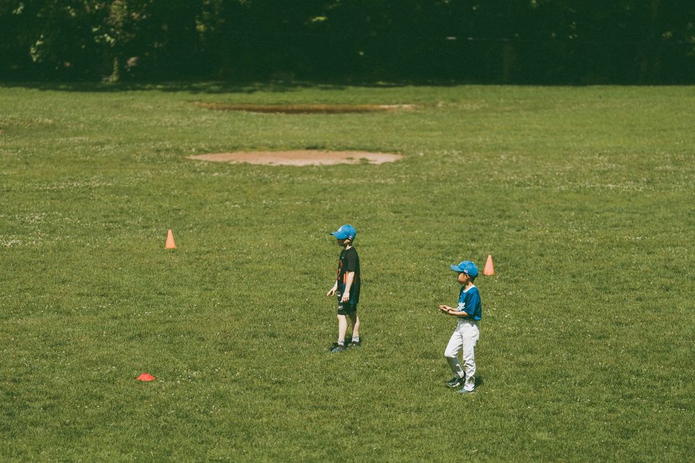 Two young baseball players in blue helmets practice passing drills on a green field with orange cones.